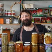 Comerciante sorrindo atrás de prateleiras com conservas em uma pequena loja.
