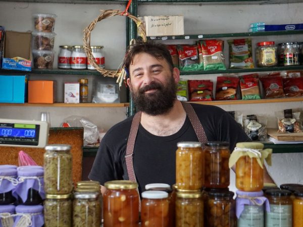 Comerciante sorrindo atrás de prateleiras com conservas em uma pequena loja.