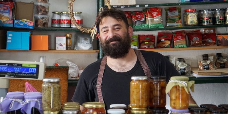 Comerciante sorrindo atrás de prateleiras com conservas em uma pequena loja.